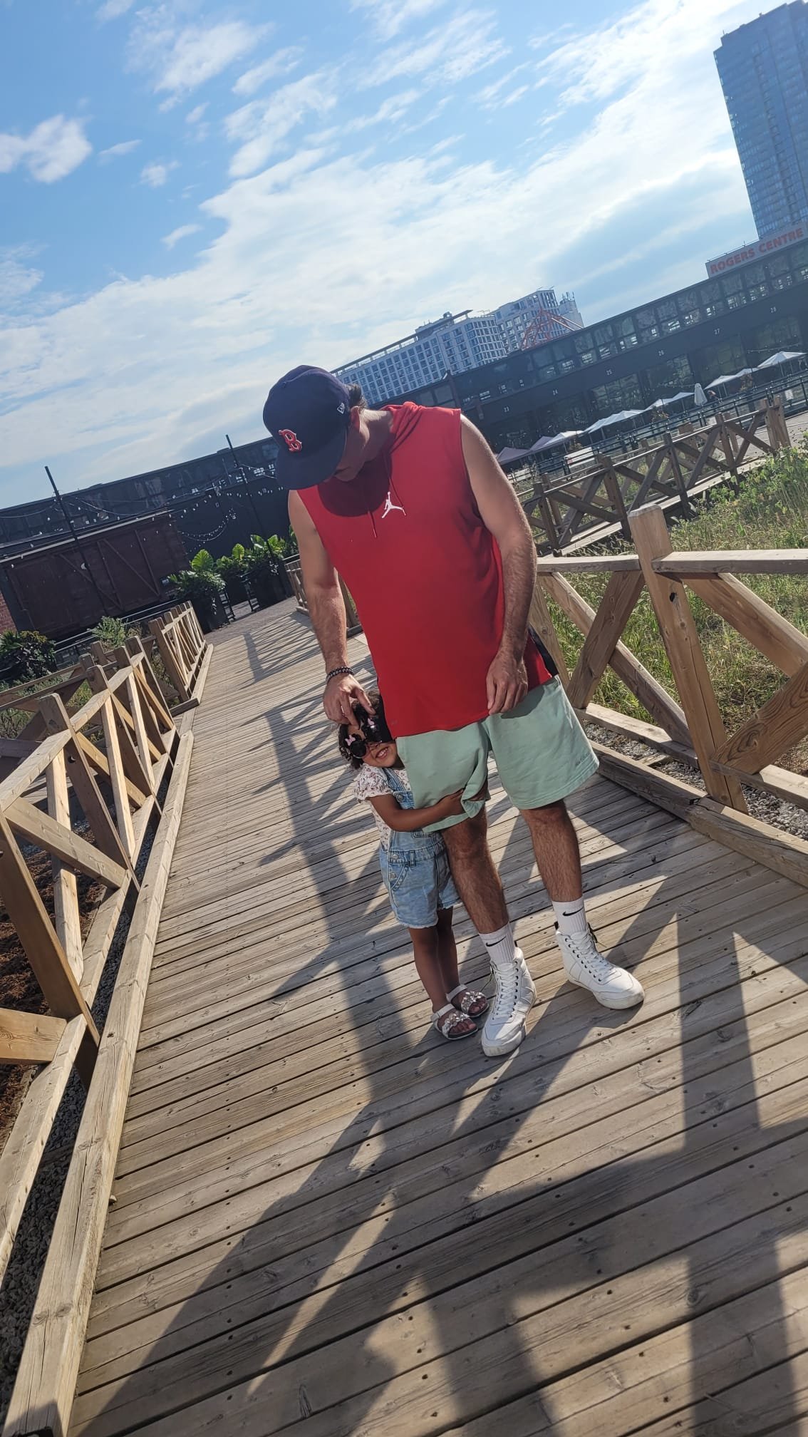 Man in red shirt and cap on a wooden rooftop deck with his young daughter hugging his leg, city buildings and blue sky behind them